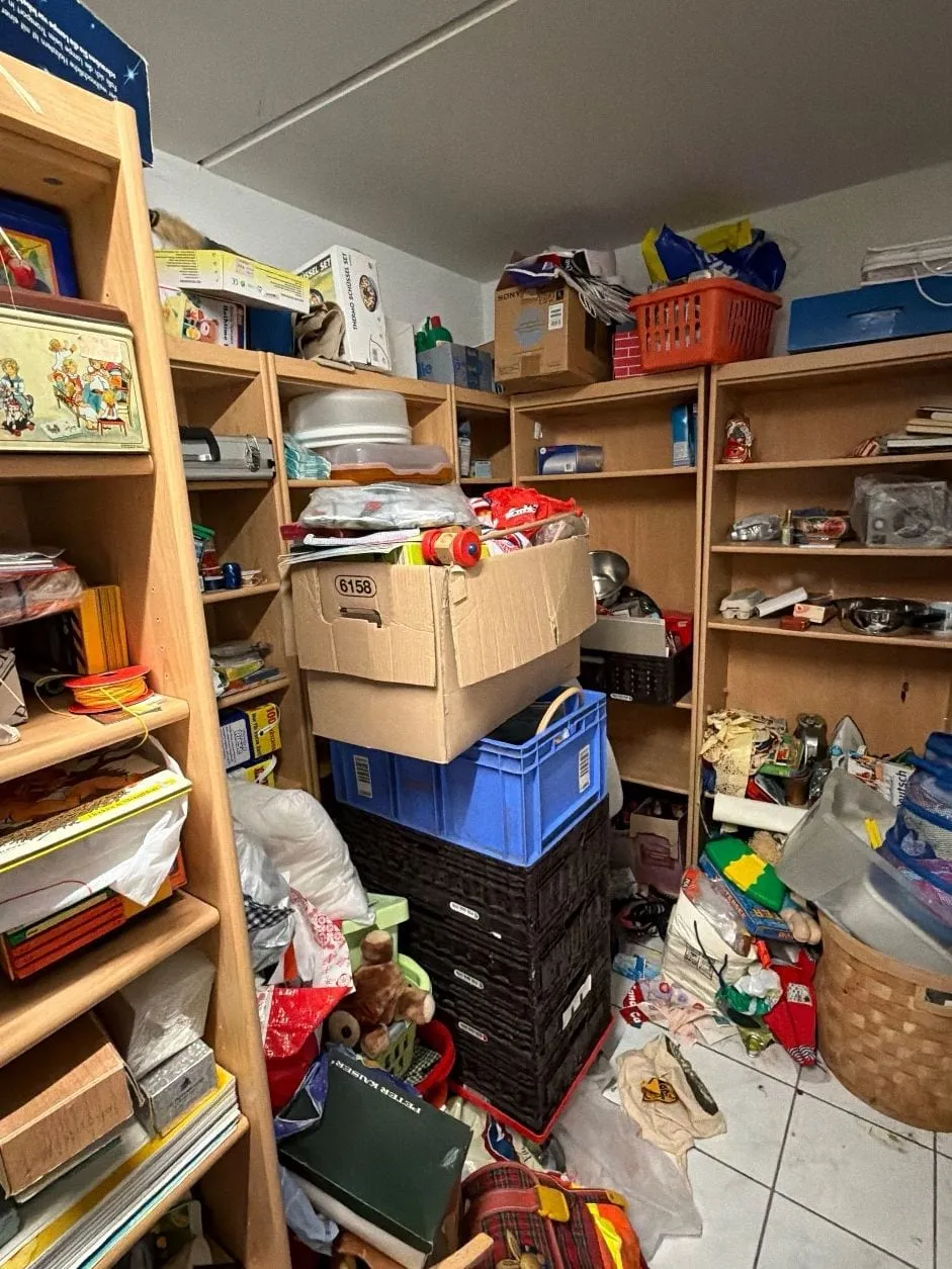 Kellerraum mit Holzregalen an den Wänden, voll mit Kartons, Dosen und Haushaltsgegenständen, davor gestapelte Kisten, Körbe und Taschen auf dem Fliesenboden.