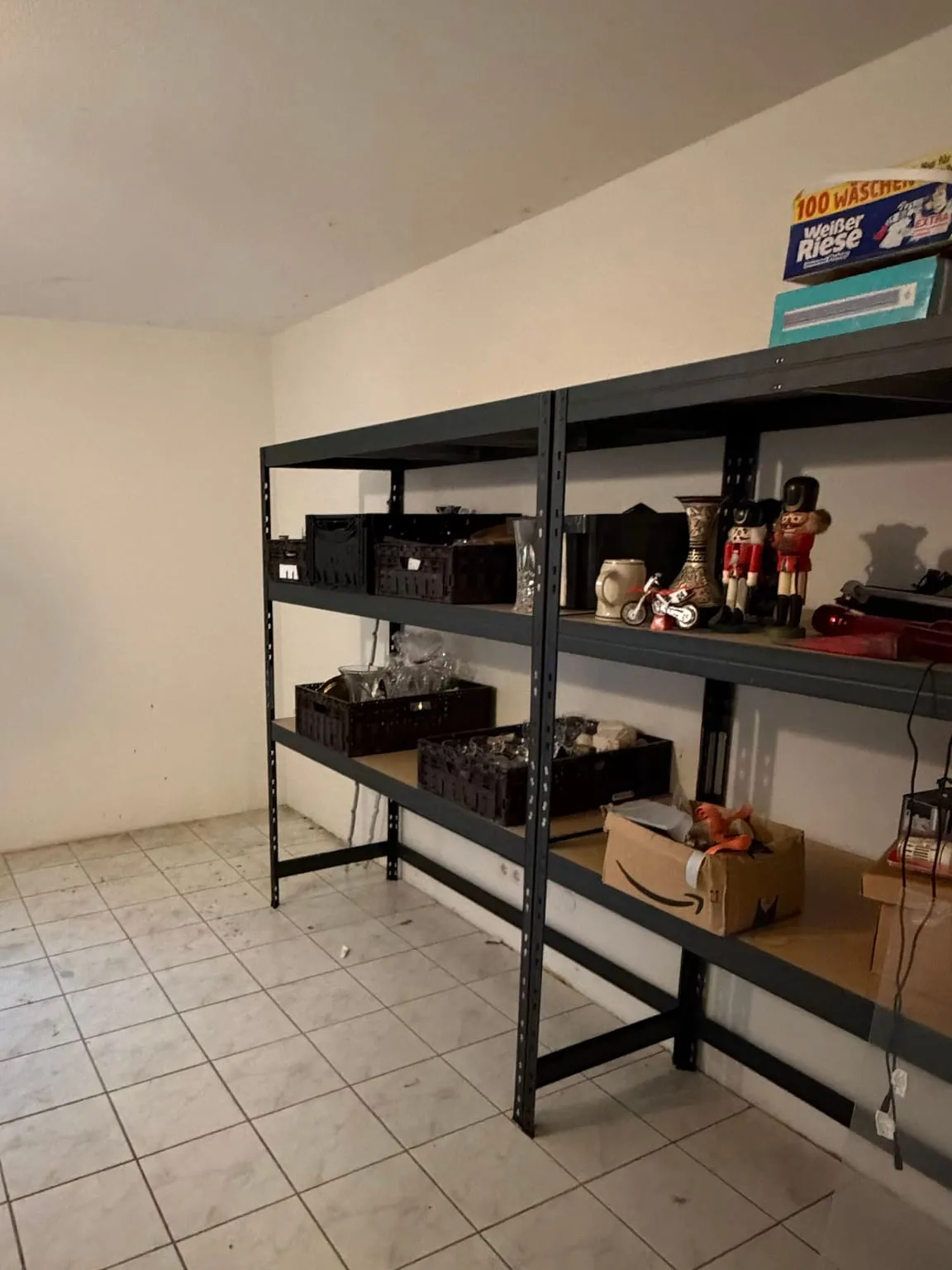 Dasselbe Metallregal schräg von der Seite, mit nur noch wenigen Körben und Kartons, davor ein freier weißer Fliesenboden.