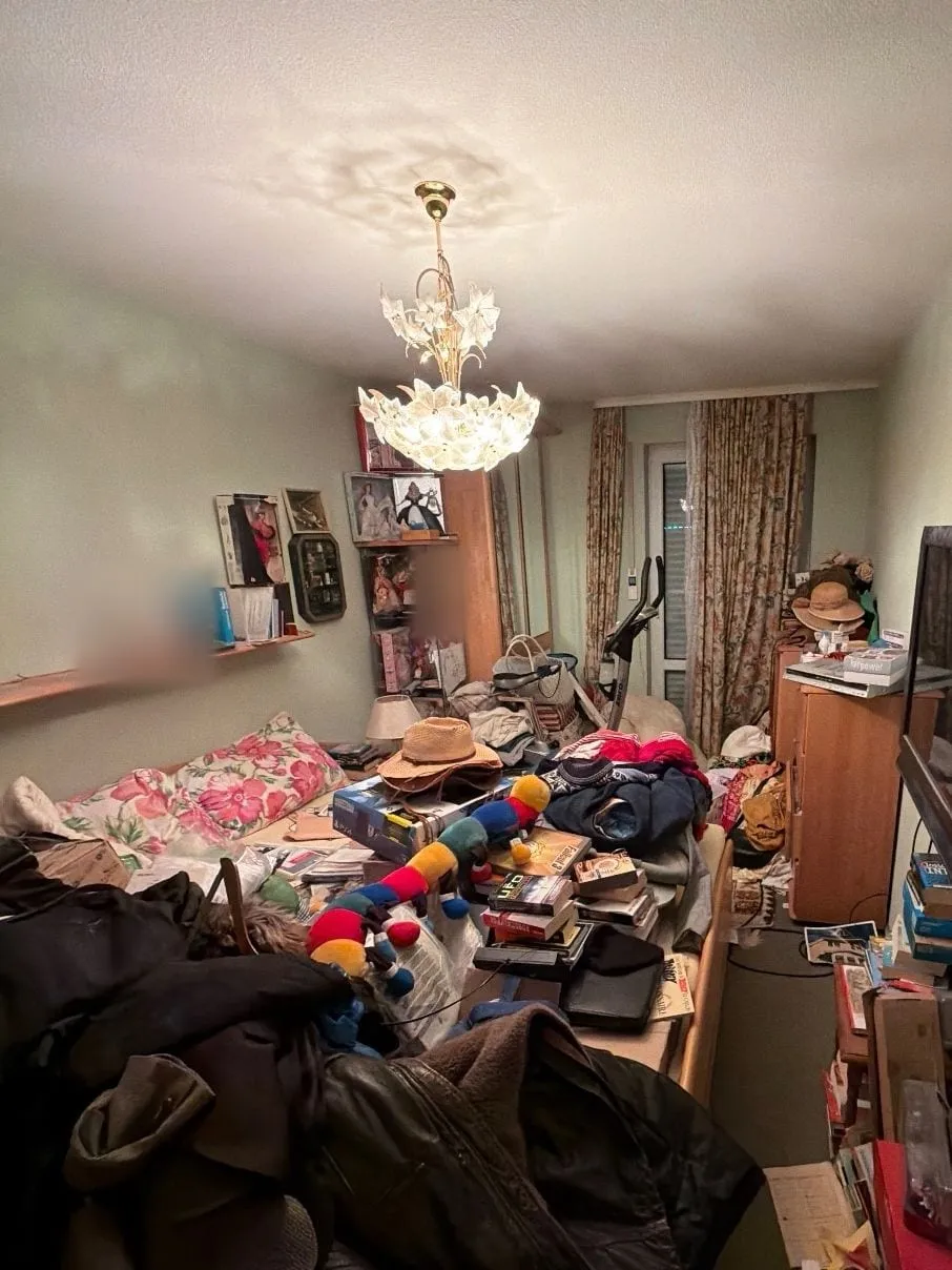 Zimmer mit mintgrünen Wänden und einem Deckenleuchter mit weißen Blüten, Bett und Boden sind mit Kleidung, Büchern und Kartons bedeckt.