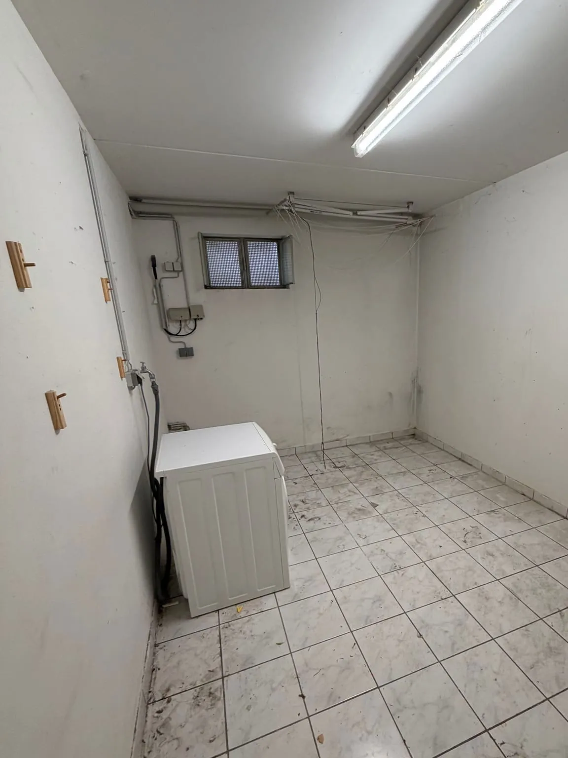 Derselbe Kellerraum geräumt, mit freiem Fliesenboden, leeren Wänden und der Waschmaschine an der linken Wand.