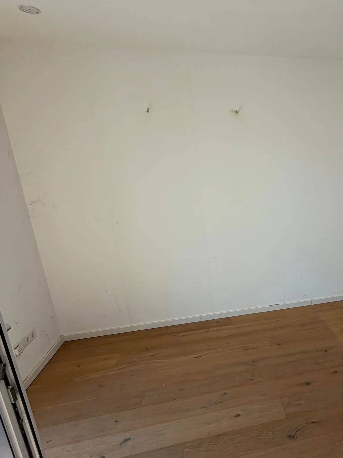 Dieselbe Raumecke nach der Räumung mit leerer weißer Wand, zwei Haken und freiem Holzboden, links unten der Rahmen der geöffneten Balkontür.