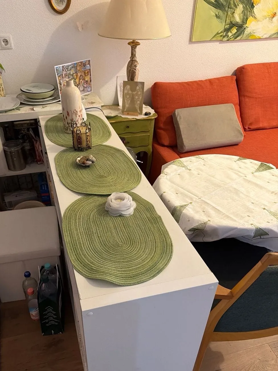 Weißes Sideboard mit grünen Tischsets als Raumteiler, dahinter ein Regal mit Geschirr, eine Tischlampe auf einer grünen Kommode, ein orangefarbenes Sofa und ein runder Tisch mit Tischdecke