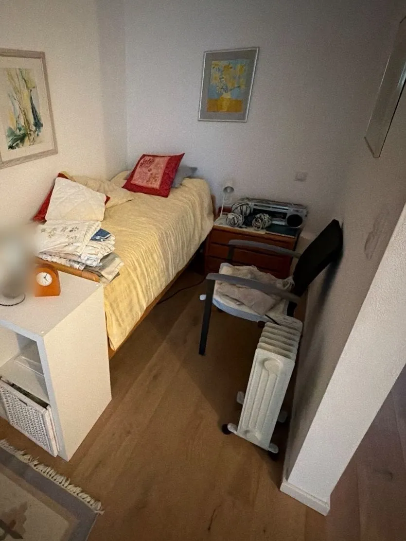 Schlafnische mit Einzelbett, Nachttisch, Stuhl, Ölradiator und Würfelregal, an den Wänden zwei gerahmte Bilder