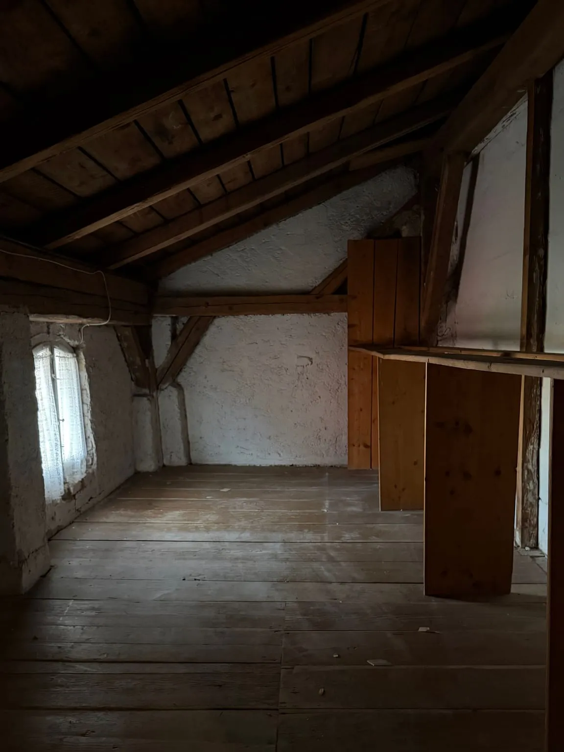 Leerer Dachboden mit freiem Dielenboden, weiß verputzter Giebelwand, kleinem Rundbogenfenster links und Holzbrettern rechts.