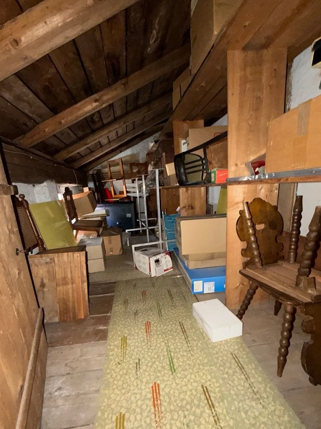Dachboden unter der Holzschalung mit Regalen voller Kartons, Holzstühlen, Platten und einem grünen Läufer auf dem Dielenboden.