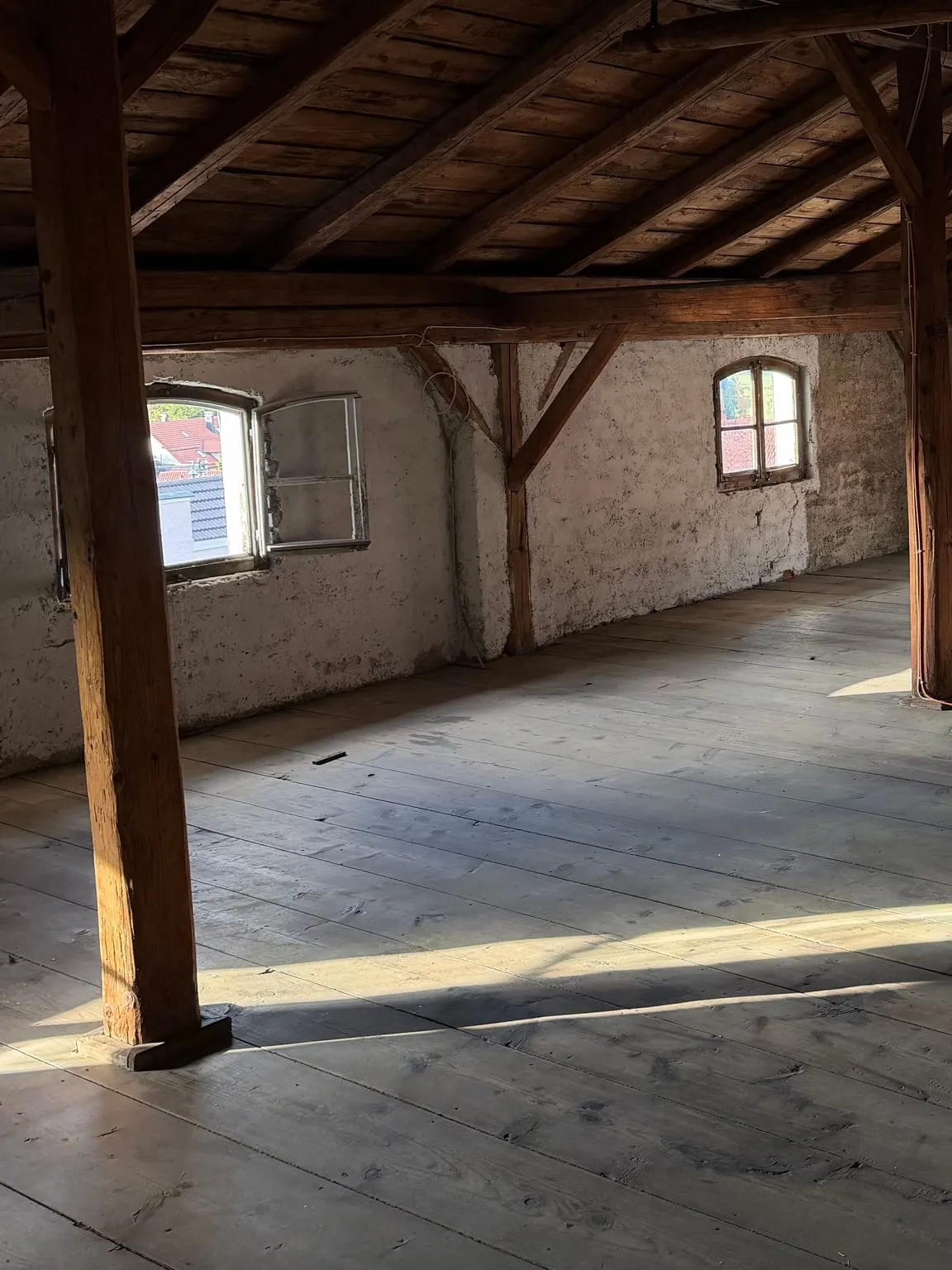 Leerer Dachboden mit blanken Holzdielen, offenem Dachstuhl und zwei Bogenfenstern in der verputzten Wand.