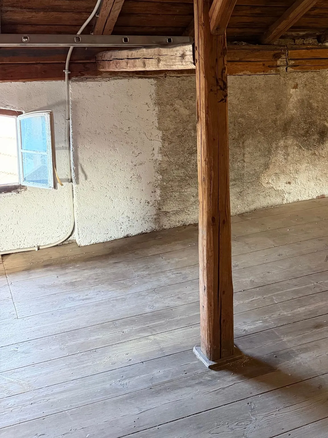 Leerer Dachboden mit Holzdielen, einem Stützpfosten, teils geweisster, teils grauer Wand und geöffnetem Fensterflügel links.