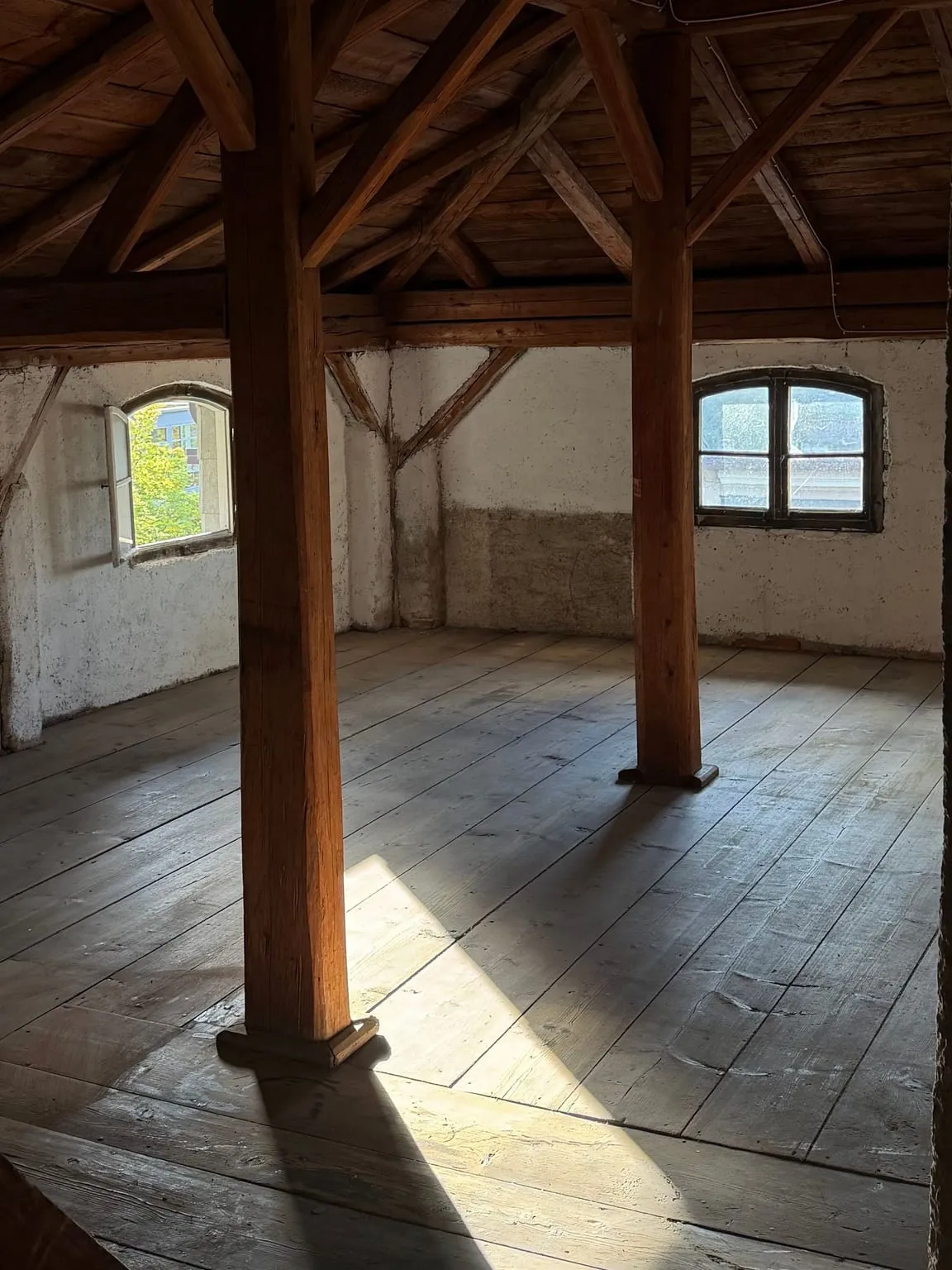 Leerer Dachboden mit freiem Bretterboden, Holzständern, Dachbalken und einem Bogenfenster in der verputzten Wand.