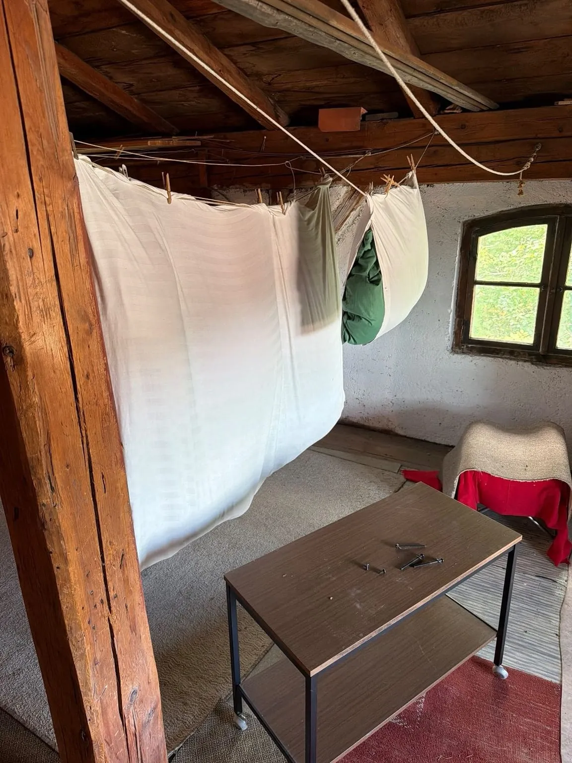 Dachboden mit Holzständer, aufgehängten Laken, einem Beistelltisch, einem mit Stoff abgedeckten Hocker und Teppichen vor einem Bogenfenster.