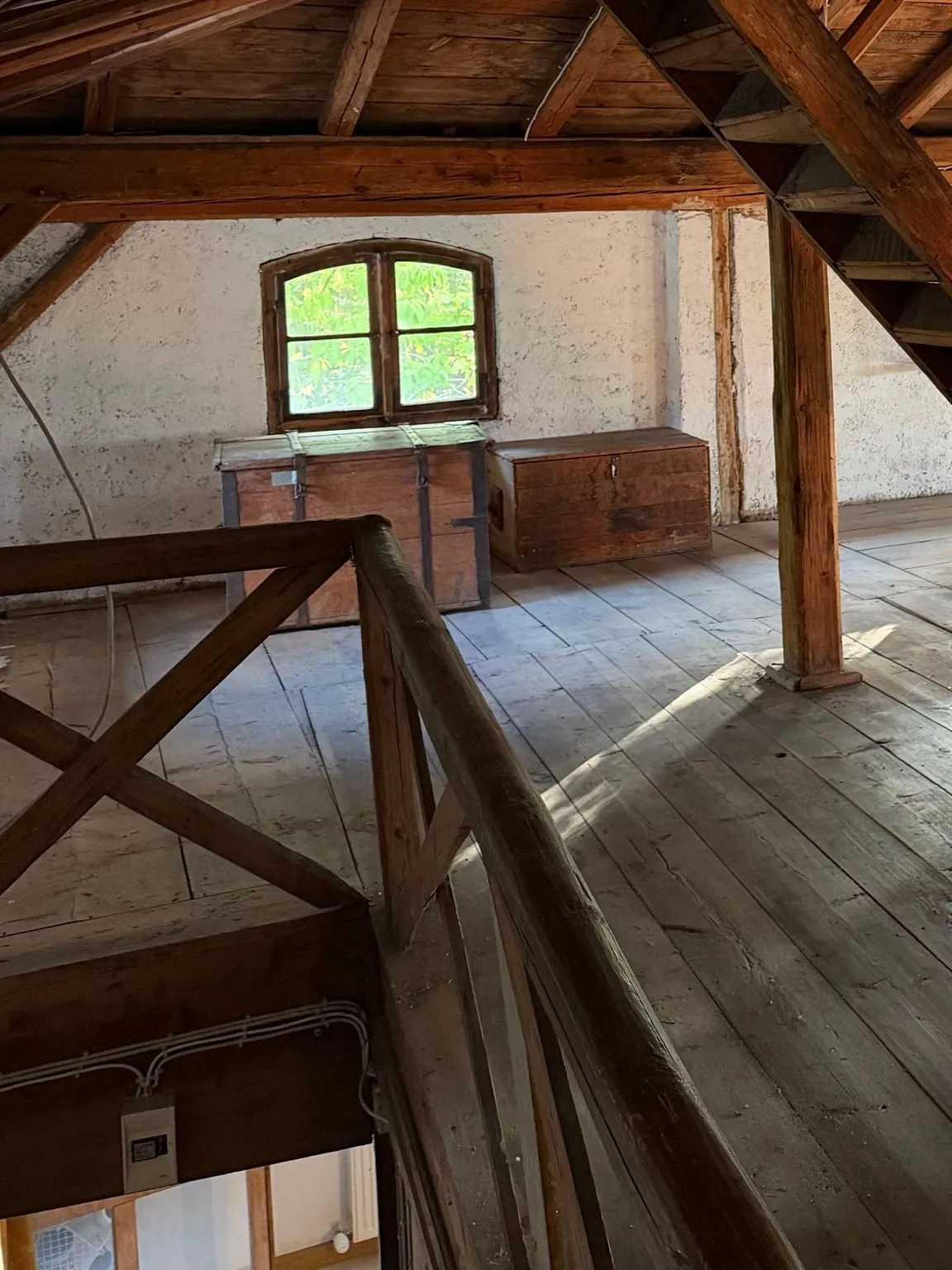 Geräumter Dachboden mit Holzdielen und Dachbalken, unter einem Bogenfenster stehen zwei Holztruhen.