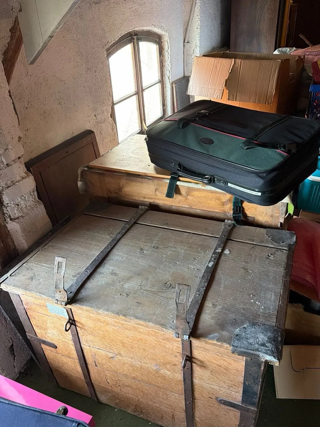 Holztruhe mit Eisenbeschlägen unter einem Bogenfenster auf einem Dachboden, umgeben von Koffern, Kartons und Kunststoffkisten.