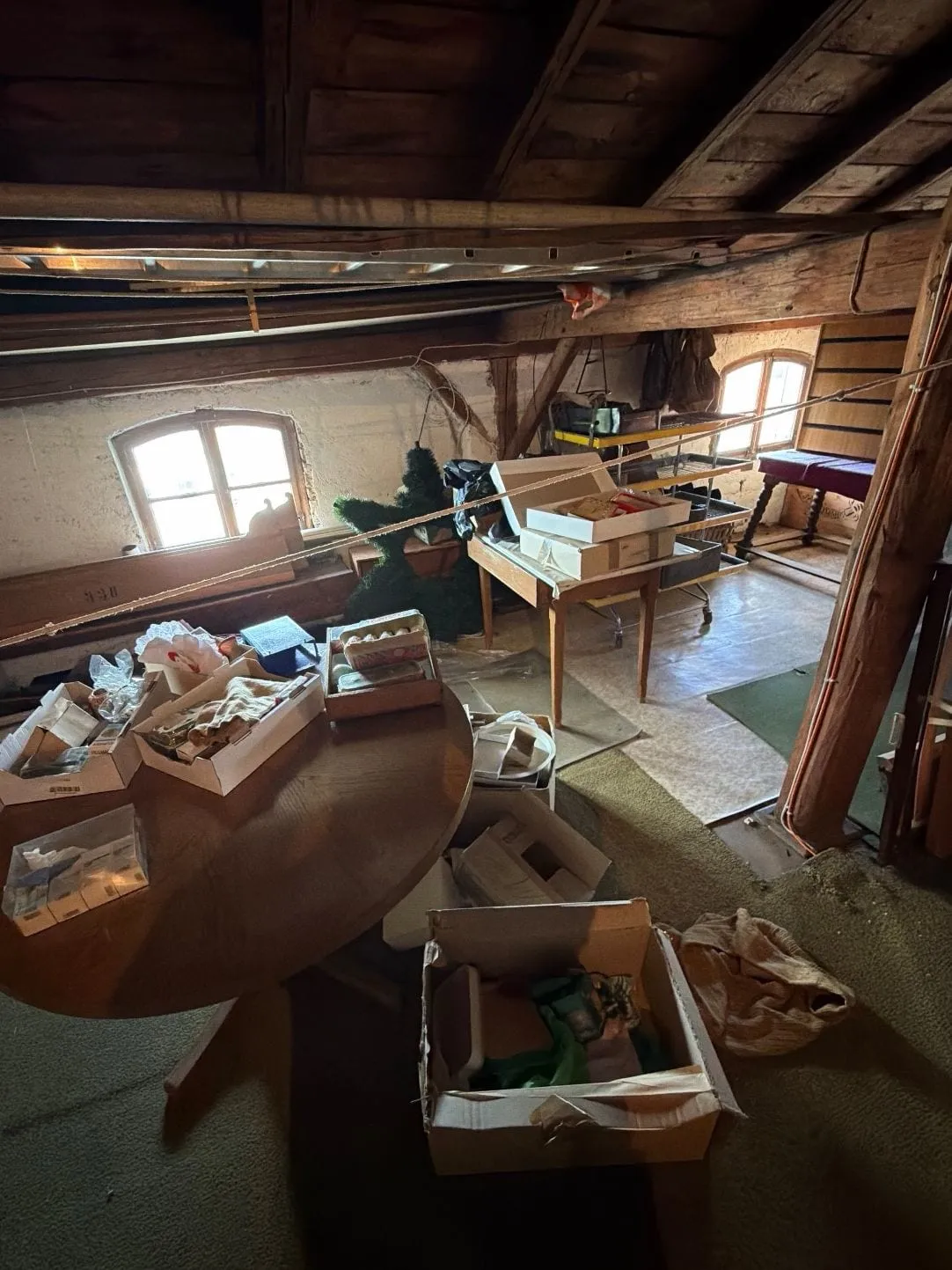 Dachboden mit Holzbalken, zwei Bogenfenstern, einem runden Tisch voller Kartons, einem Regalwagen und Teppichbahnen auf dem Boden.