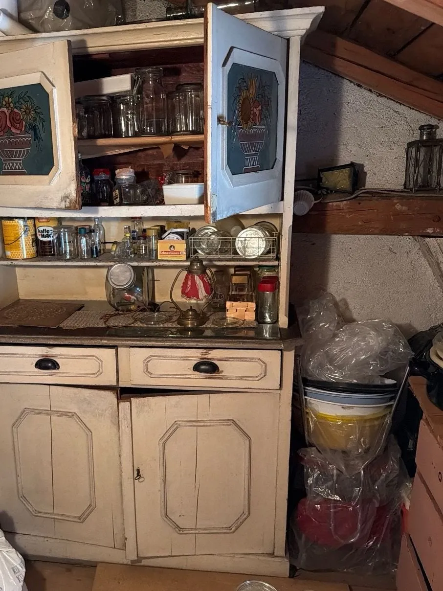 Dachkammer vor der Räumung: helles Küchenbuffet mit bemalten Türen und Einmachgläsern, daneben Plastiksäcke mit Schüsseln und eine rosa Kommode unter einem Holzbalken mit Laternen