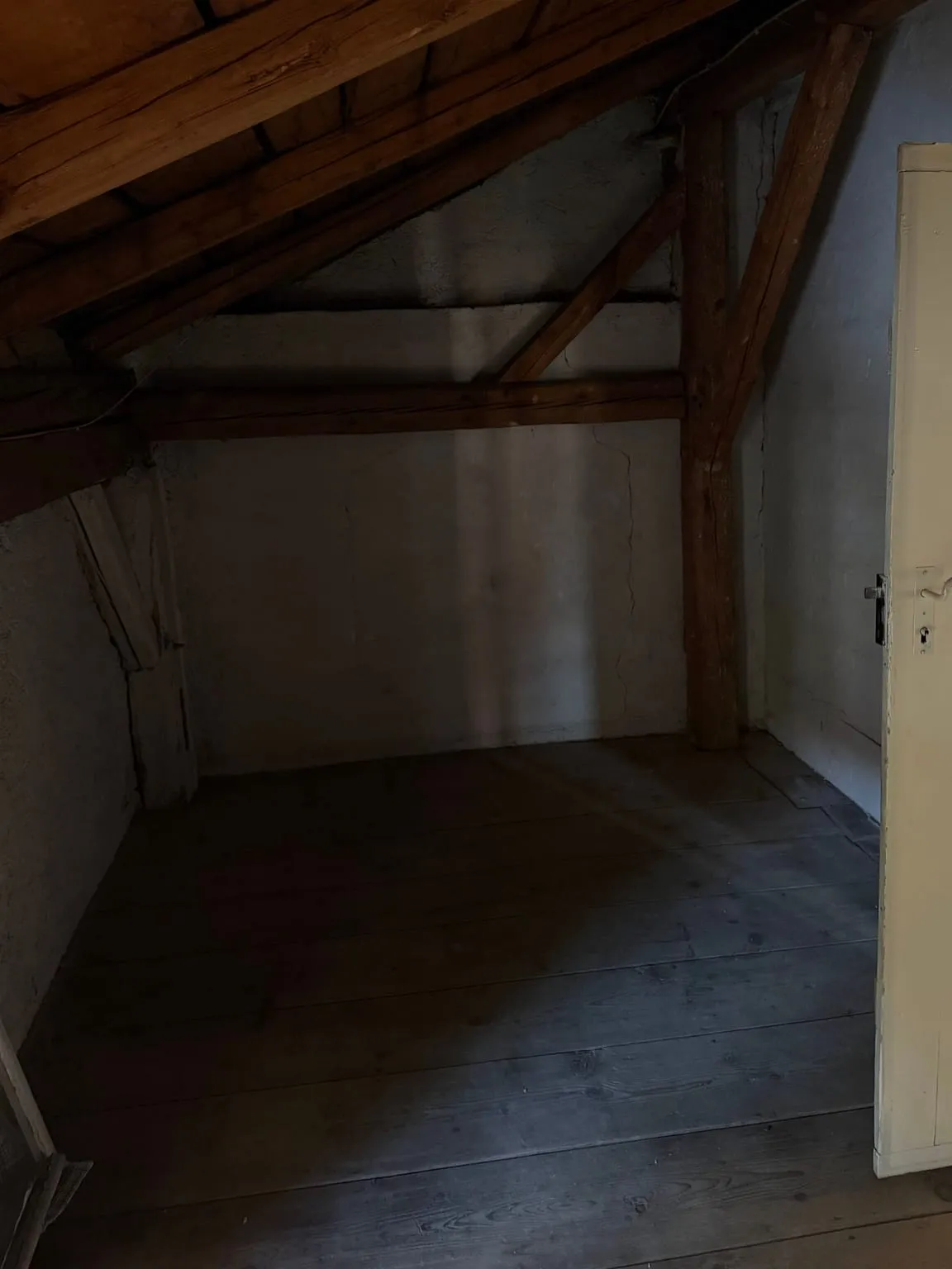Dieselbe Dachkammer leer geräumt, mit freiem Holzdielenboden, weiß verputzter Wand, Holzpfosten und geöffneter weißer Tür.