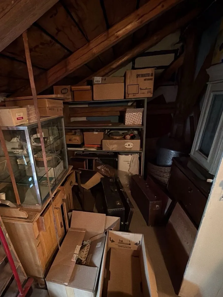 Dachkammer unter Holzsparren mit einem Metallregal voller Kartons, einer Glasvitrine auf einem Holzschrank, Koffern und offenen Kartons auf dem Boden.