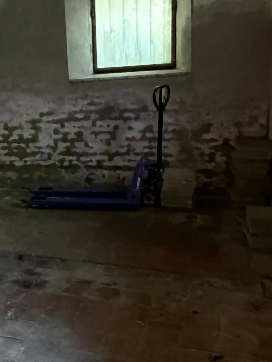 Dieselbe Wand im Gewölberaum mit Fenster und weißer Laibung, davor ein Hubwagen und ein Plattenstapel auf dem Ziegelboden