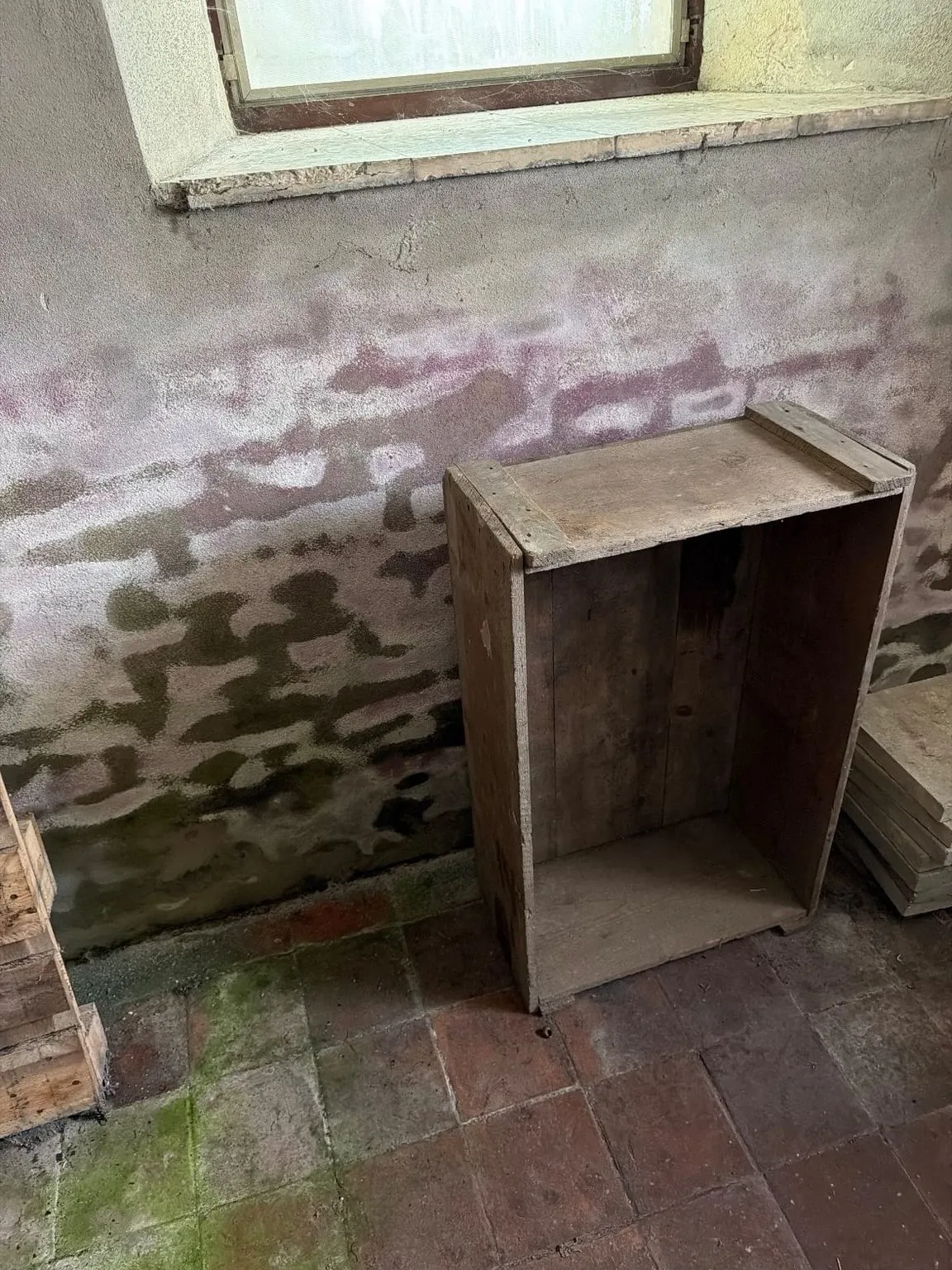 Leere Holzkiste hochkant vor einer fleckigen Wand unter einem Fenster mit breitem Sims, links eine Palette, rechts gestapelte Platten auf Ziegelboden
