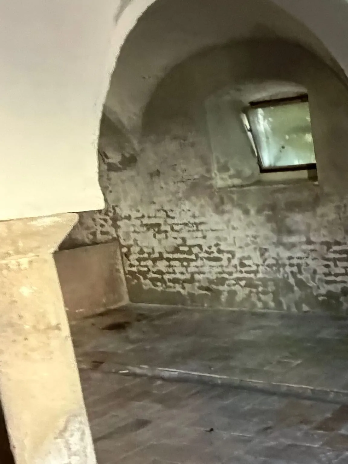 Derselbe Gewölbejoch nach der Räumung: leere Wand mit Fleckenmuster unter dem gekippten Nischenfenster, links die Steinsäule und das Ende des Betonbeckens, freier Ziegelboden