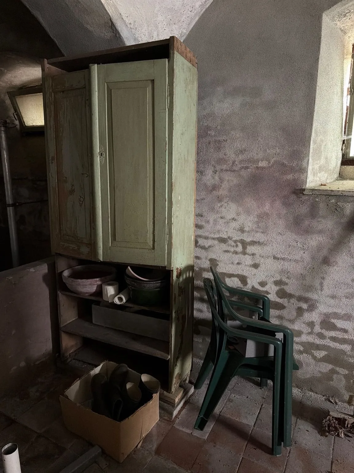 Gewölbekeller vor der Räumung: grün gestrichener Holzschrank mit Schüsseln im offenen Unterteil, daneben gestapelte grüne Gartenstühle und ein Karton mit Stiefeln, rechts ein Fenster in tiefer Laibung
