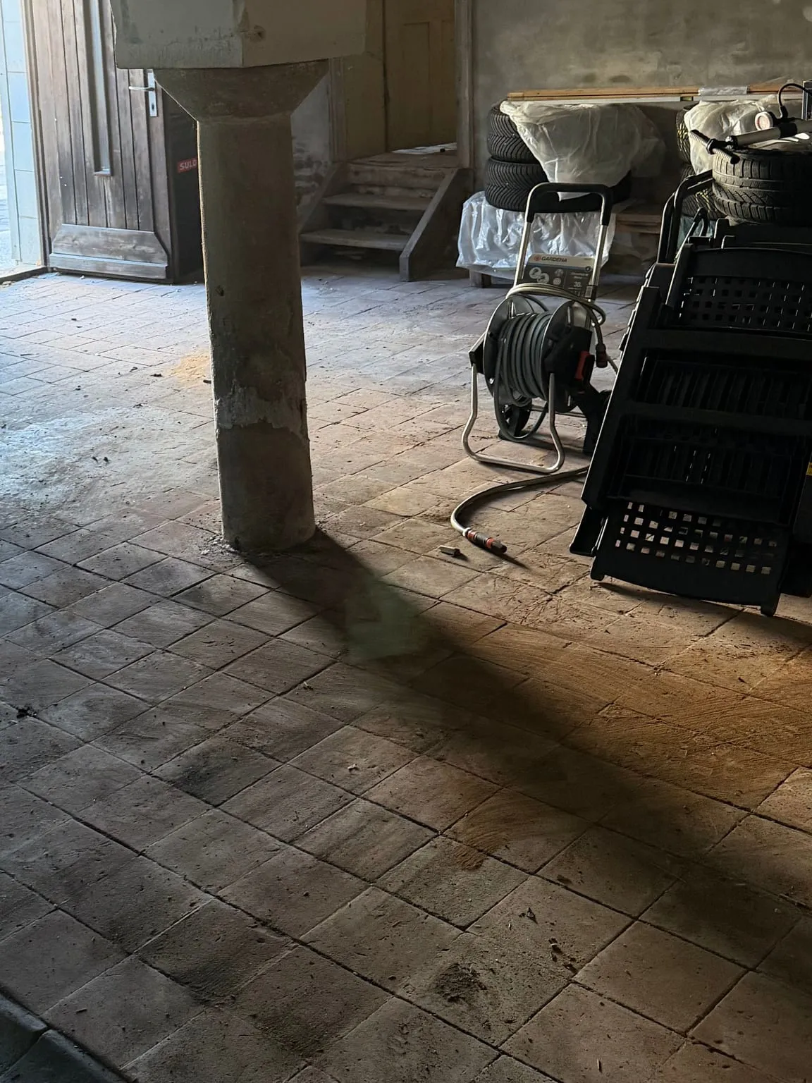 Geräumter Gewölberaum mit Steinsäule und freiem Fliesenboden, dahinter eine kleine Holztreppe, rechts Reifen, ein Schlauchwagen und schwarze Kunststoffkisten.