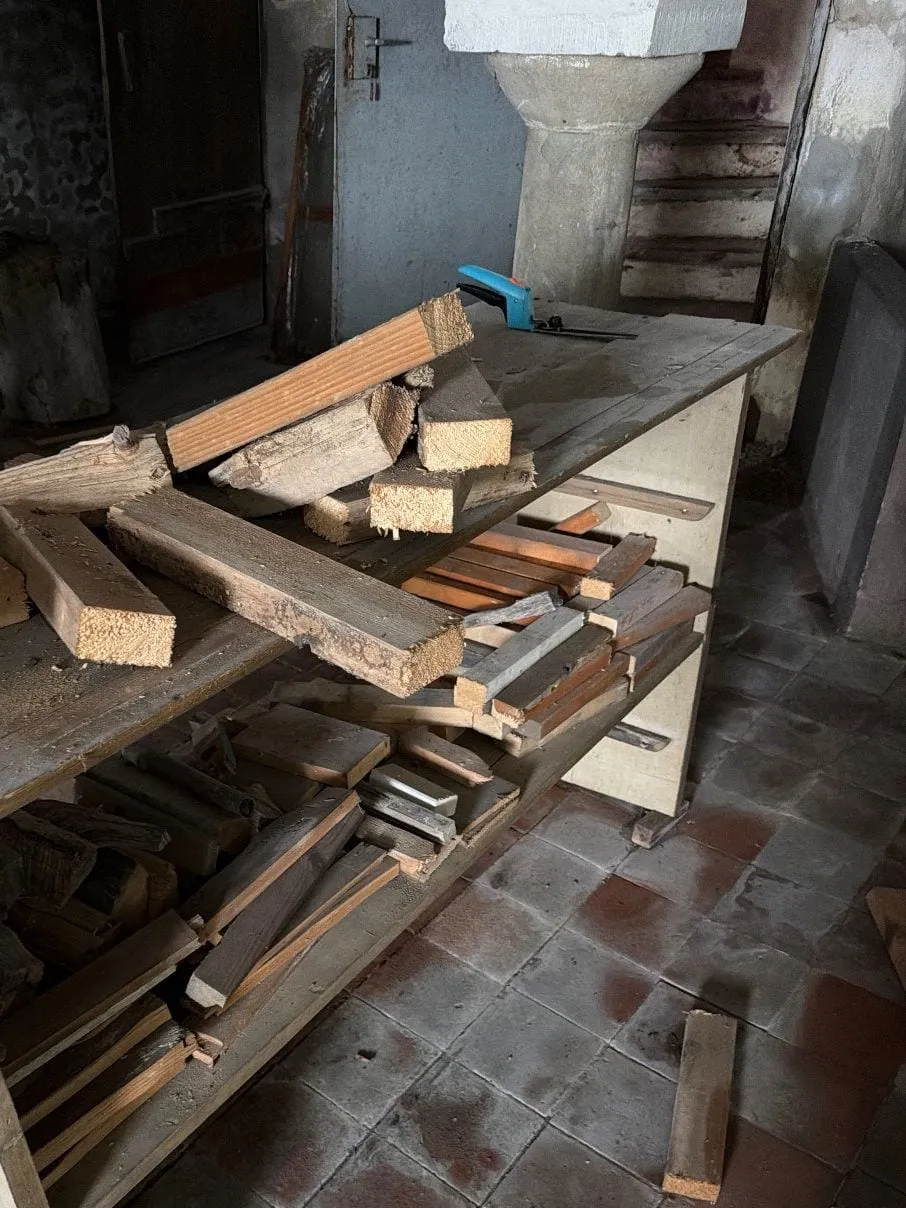 Werkbank mit gestapelten Holzlatten und Brettern vor einer Steinsäule in einem Gewölberaum mit Fliesenboden.