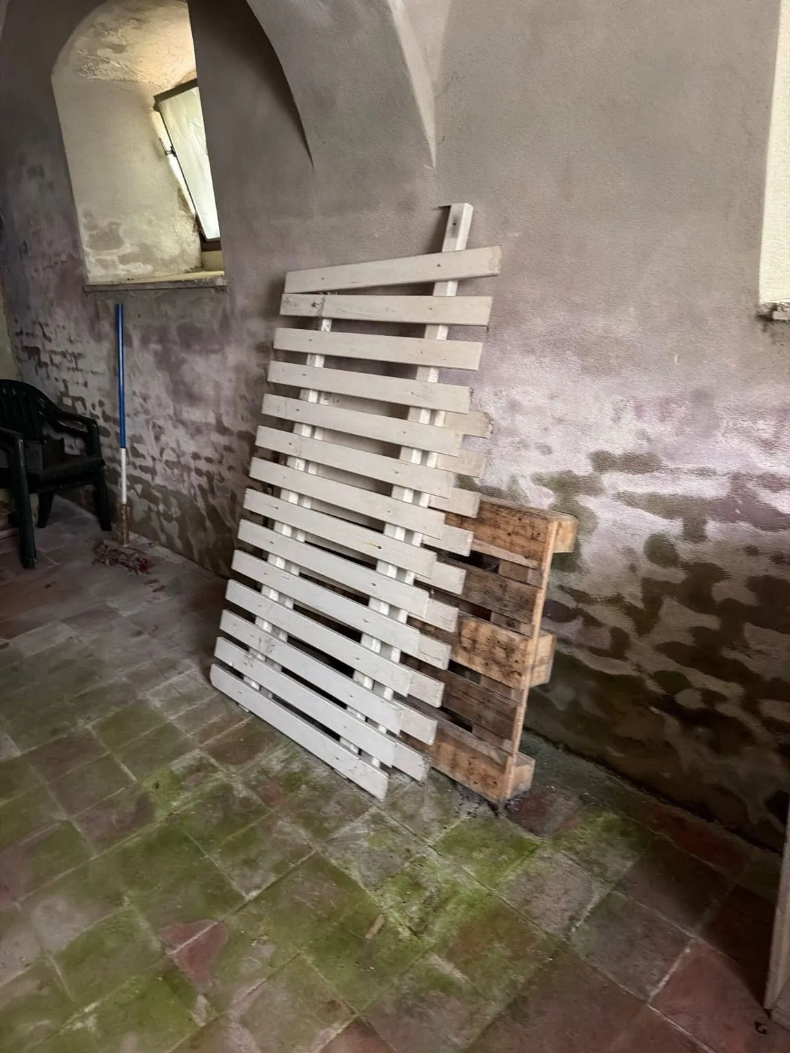 Lattenrost und Holzpalette an einer fleckigen Gewölbewand unter einer Fensternische, links ein Gartenstuhl auf dem Fliesenboden.