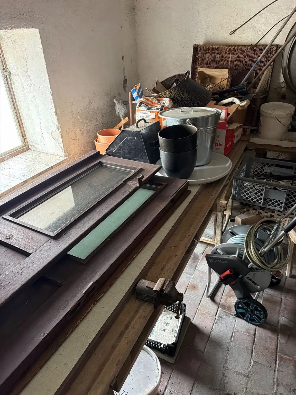 Werkbank unter einem Kellerfenster, darauf alte Fensterflügel, Töpfe, Eimer und Kisten, daneben ein Schlauchwagen auf dem Ziegelboden.