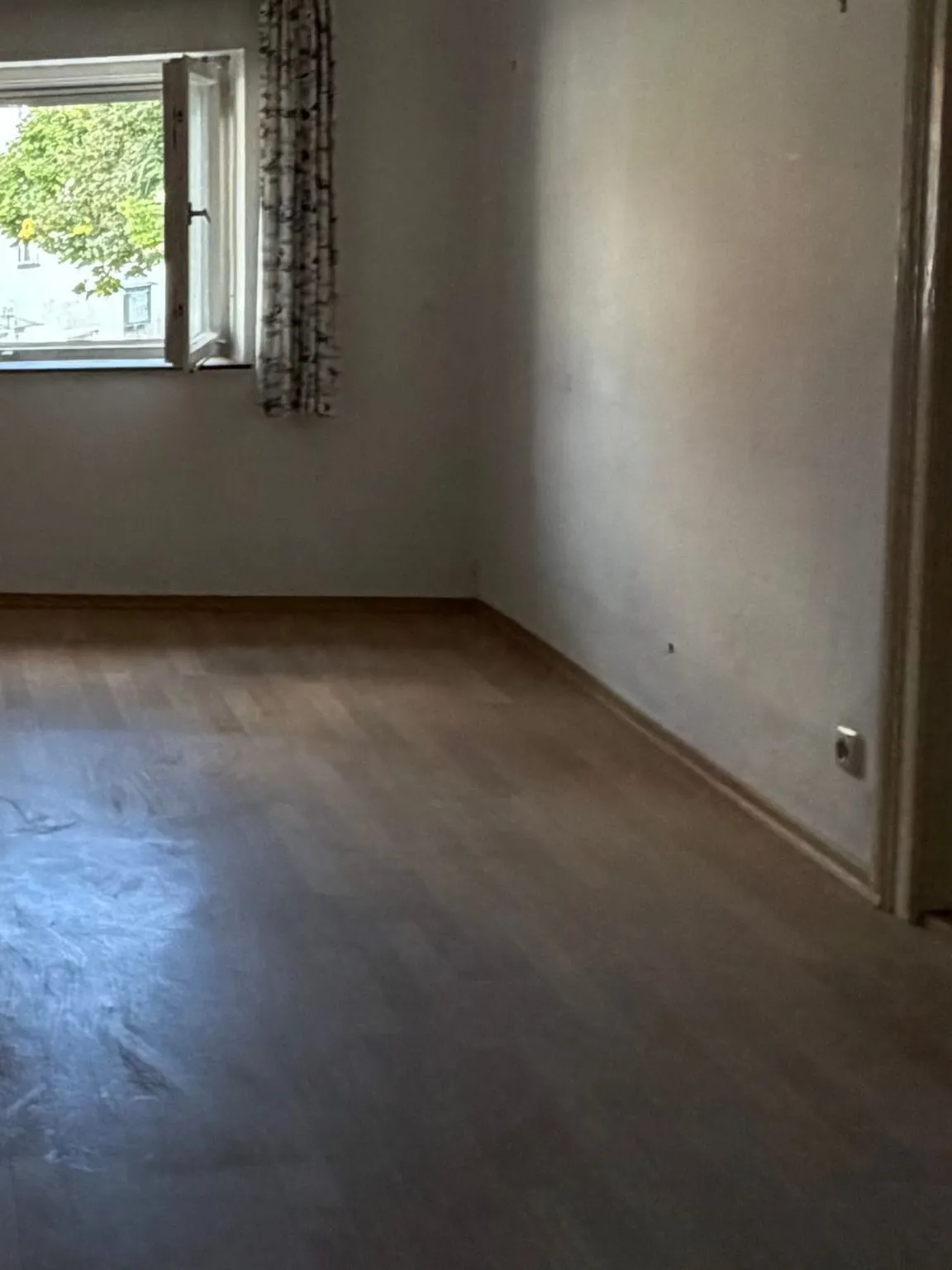 Dieselbe Schlafzimmerecke nach der Räumung mit Fenster, Vorhängen, weisser Wand und leerem Laminatboden