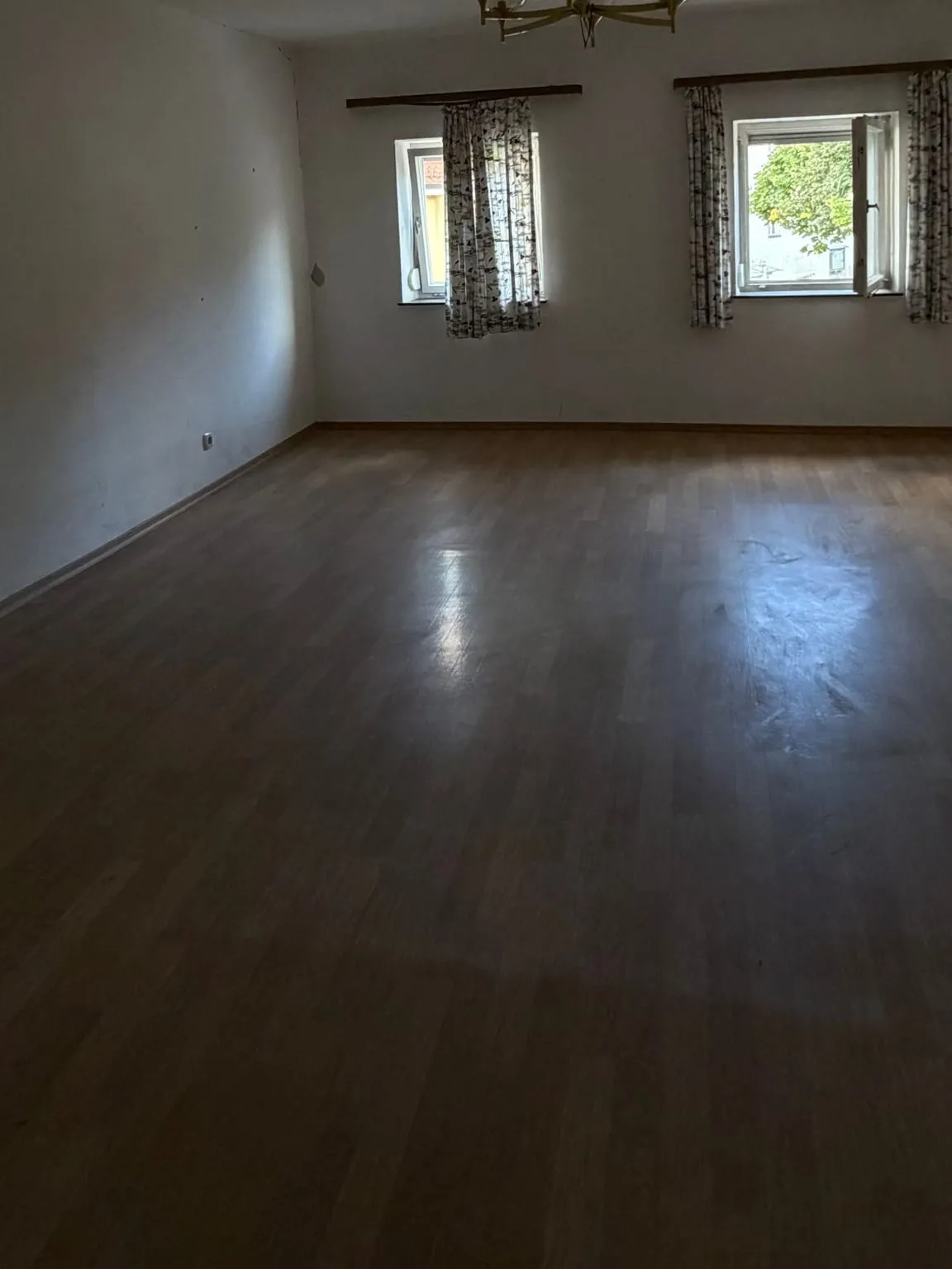 Leeres Schlafzimmer mit freiem Laminatboden und zwei Fenstern mit geblümten Vorhängen an hölzernen Vorhangleisten.