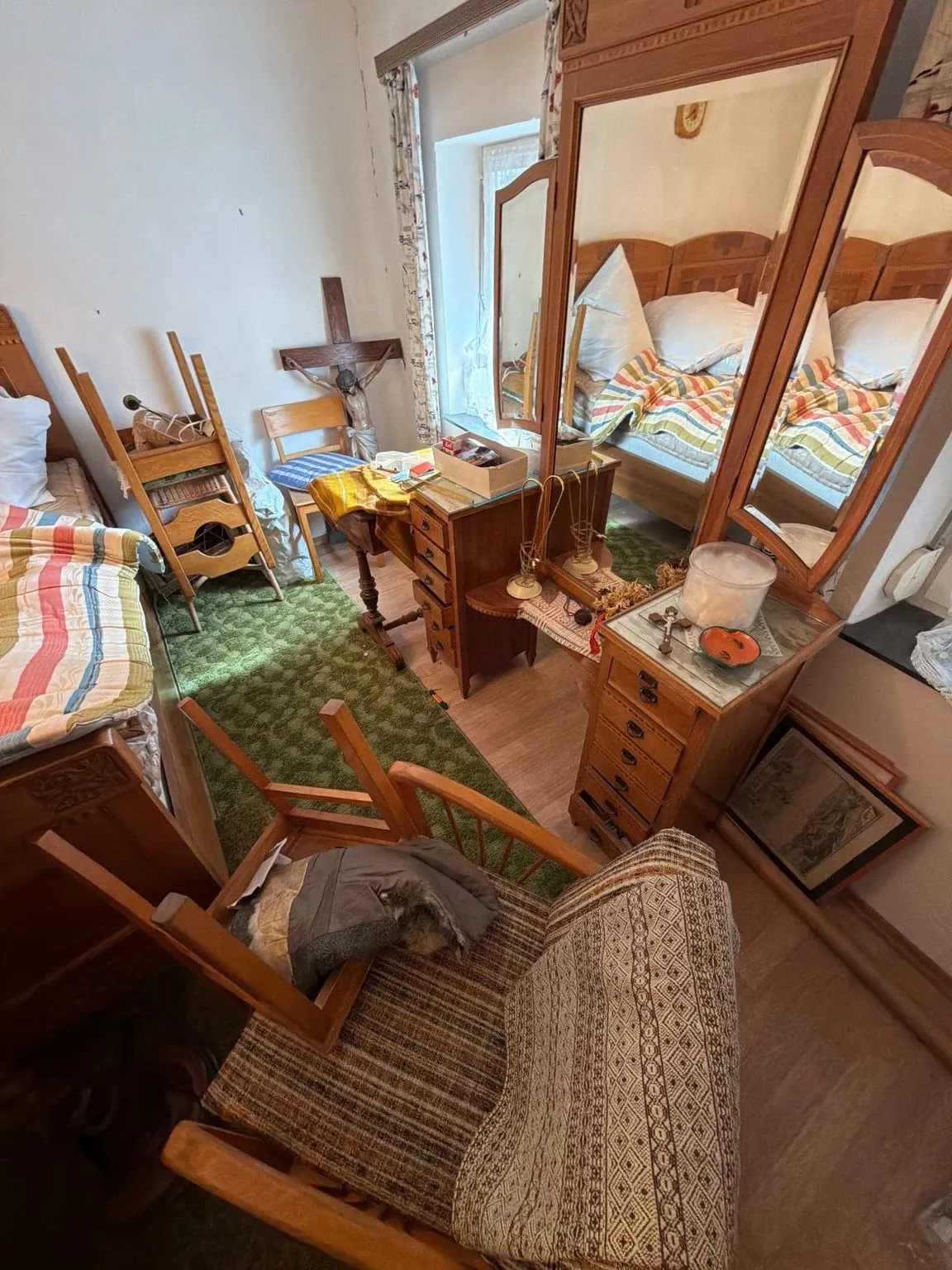 Schlafzimmer mit Betten, Spiegelkommode, Stühlen, Sessel und Kleinteilen auf grünem Teppich vor einem Fenster mit geblümten Vorhängen.