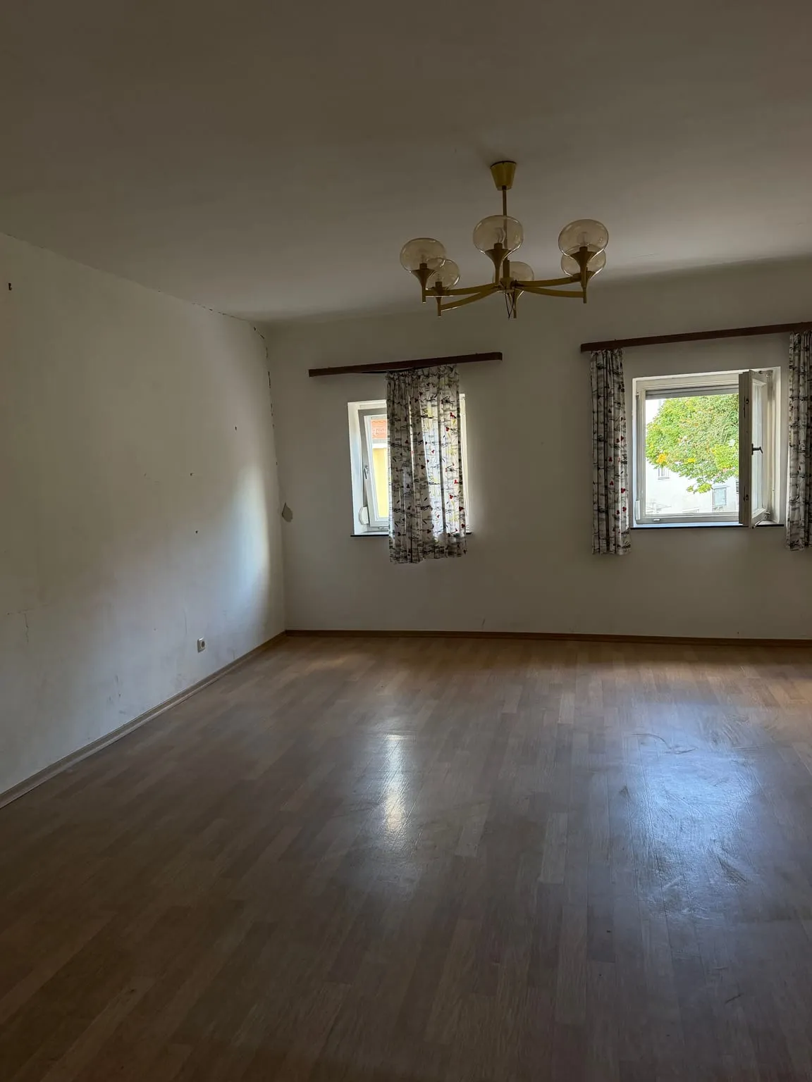 Leeres Schlafzimmer mit Laminatboden, zwei Fenstern mit gemusterten Vorhängen und dem Messing-Kronleuchter an der Decke.
