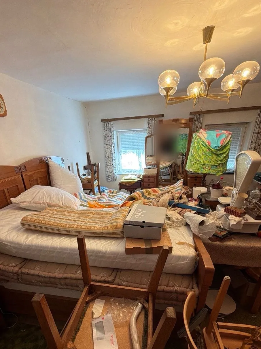 Schlafzimmer mit Doppelbett, Spiegelschrank, umgedrehten Stühlen und vielen Gegenständen auf Bett und Tisch unter einem Messing-Kronleuchter.