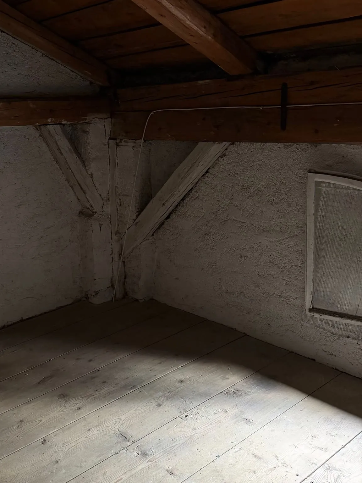 Leere Dachkammer mit Holzbalken, weiß verputzten Wänden, einem kleinen Fenster und freiem Dielenboden.
