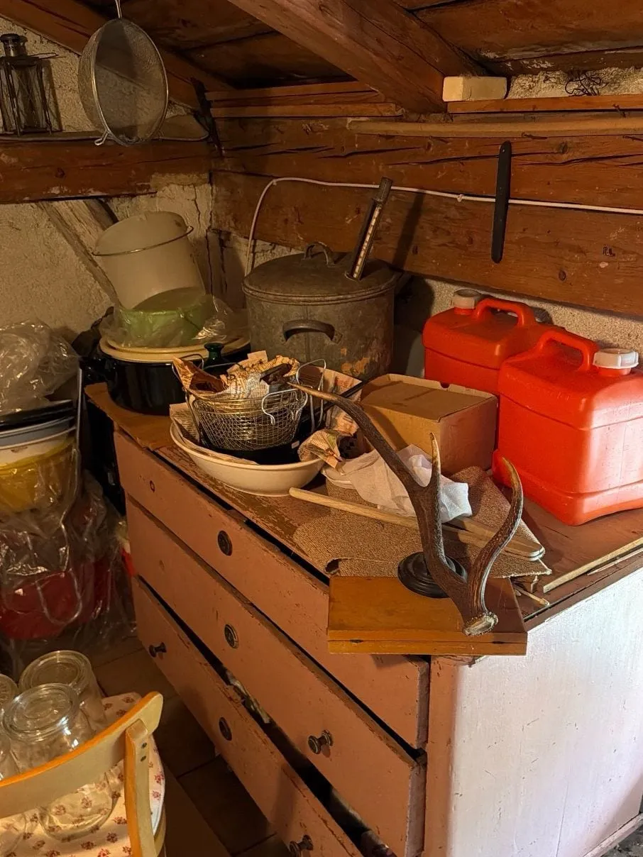 Dachkammer mit Holzbalken, davor eine rosafarbene Kommode mit Töpfen, roten Kanistern und Kartons, daneben Säcke und ein Stuhl mit Einmachgläsern.