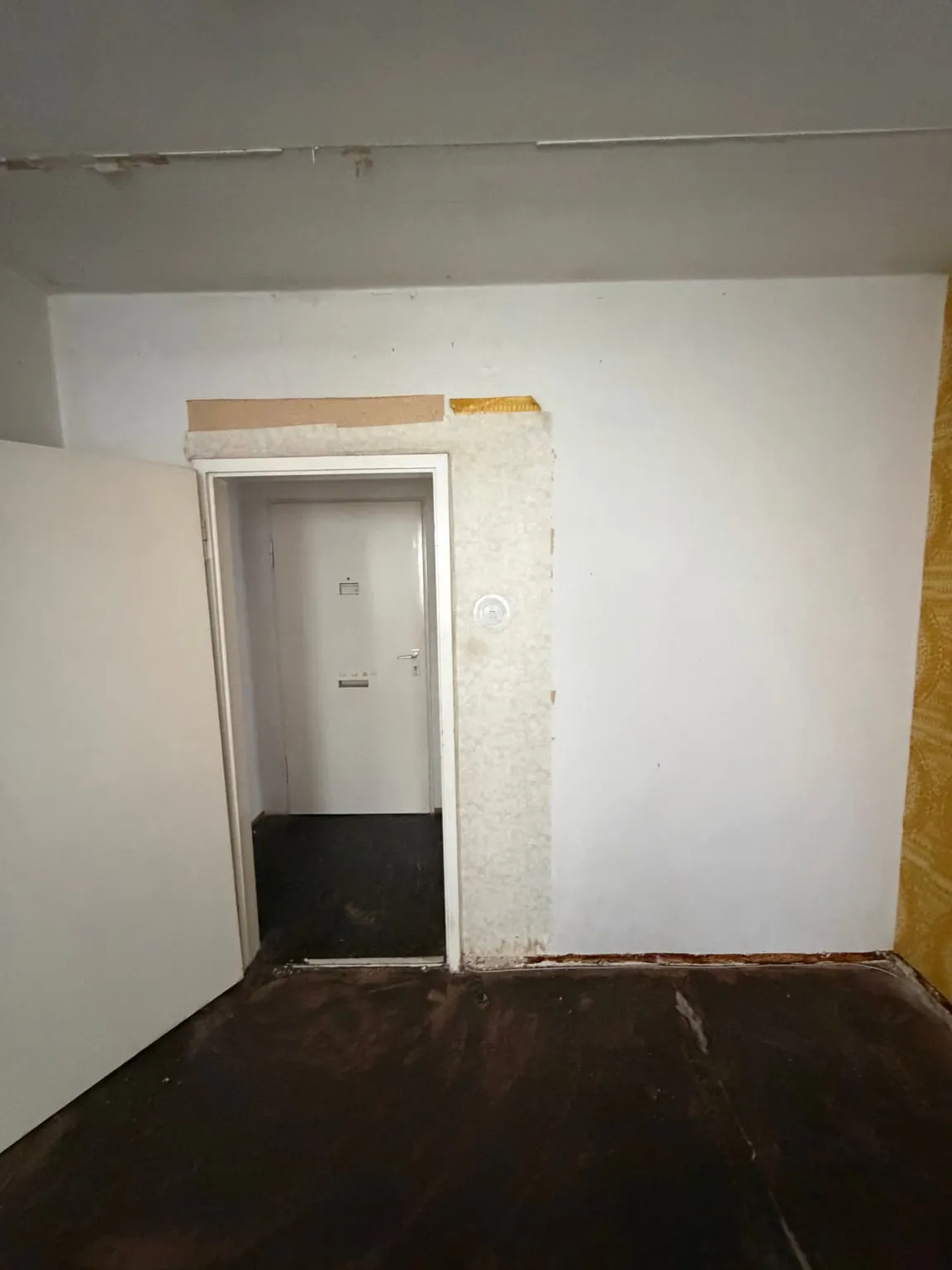 Dieselbe Wand ohne Schrank: offene Zimmertür mit Blick in den Flur, Putzreste rund um den Türrahmen, Boden ohne Teppich