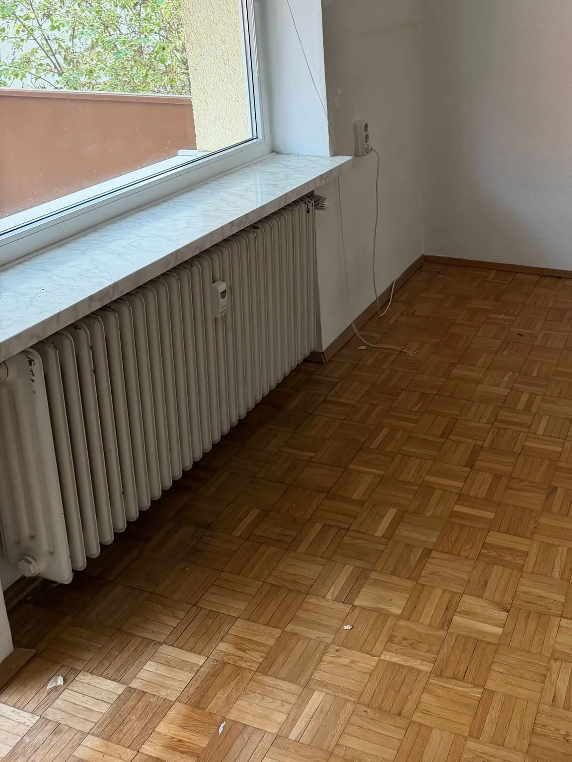 Großes Fenster mit leerer Fensterbank und Heizkörper, daneben eine freie Zimmerecke mit Parkettboden