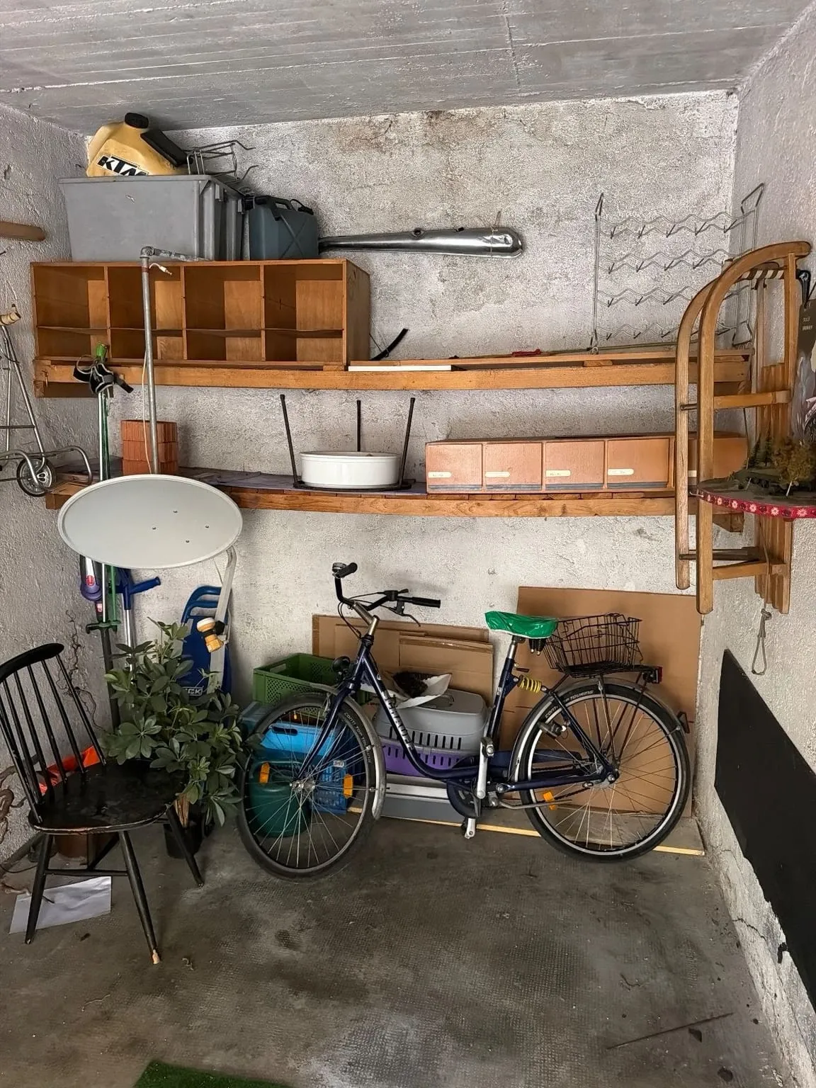Garage mit Regalbrettern voller Kisten und Kanister an der Rückwand, davor ein Fahrrad, ein Holzschlitten, ein Stuhl und Gartensachen