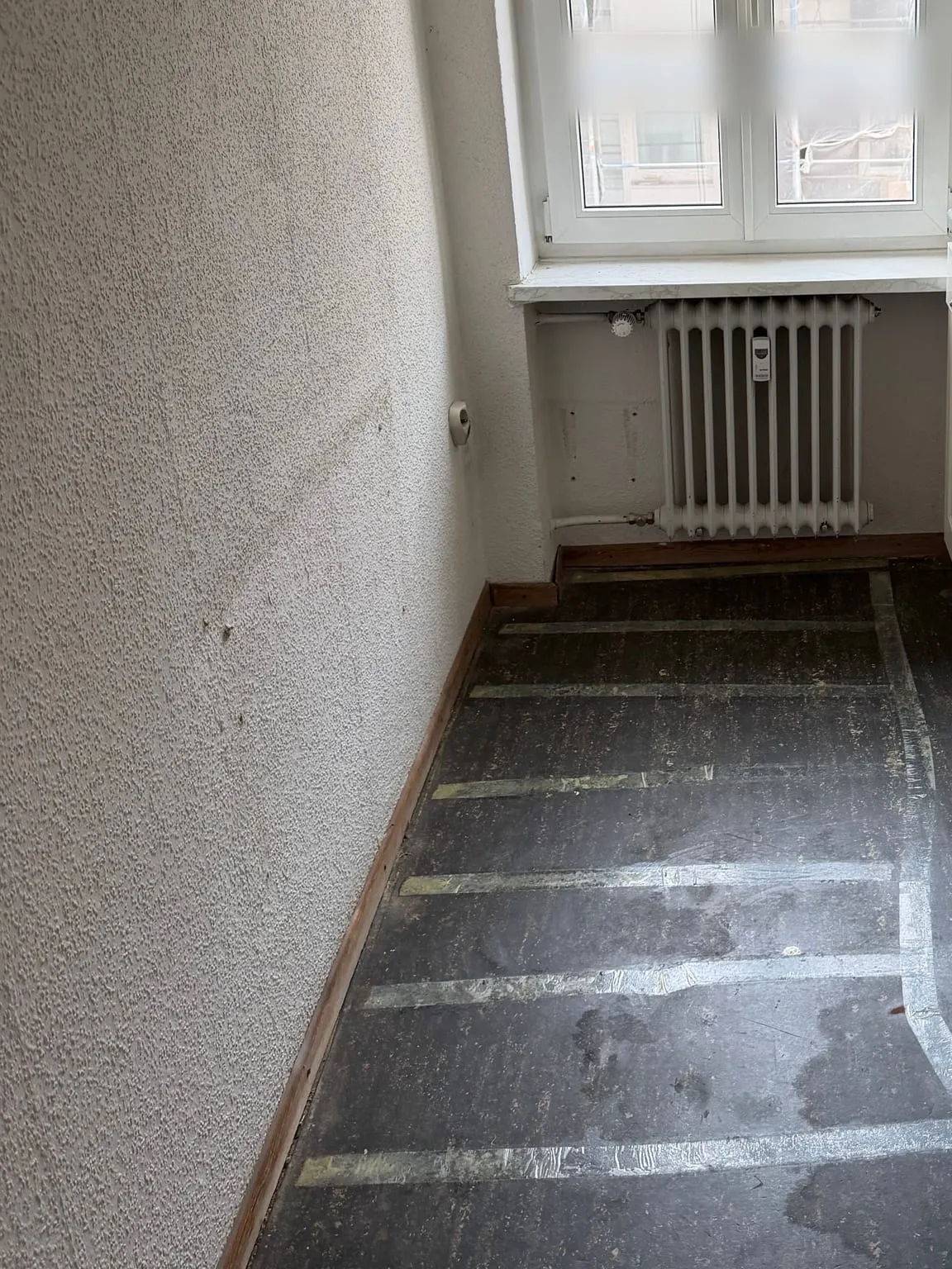 Freie Wand und Fensterseite der Küche, unter dem Fenster der Heizkörper, der Boden ohne Belag mit Klebebandresten