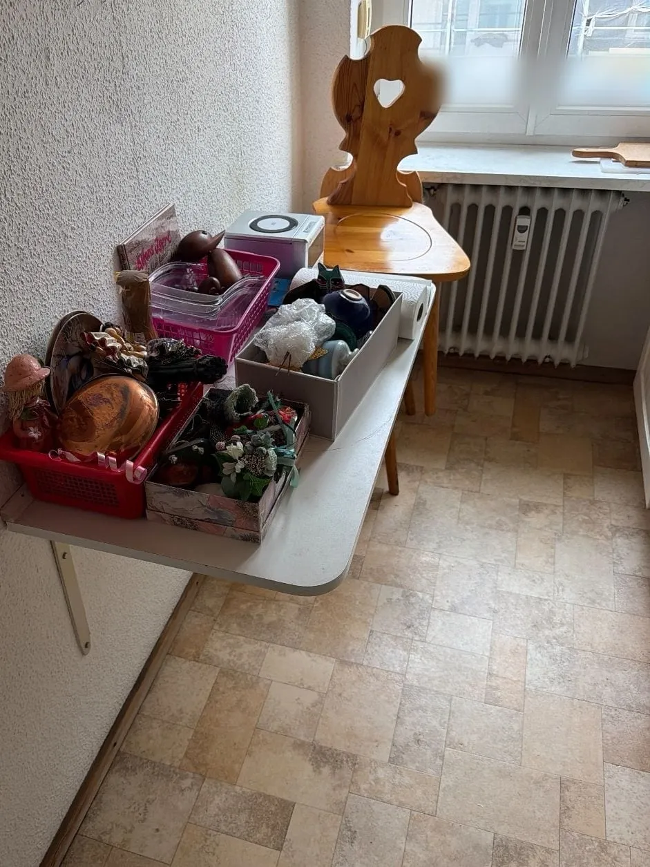 Klapptisch an der Wand voller Kisten mit Hausrat, dahinter ein Holzstuhl vor Fenster und Heizkörper, Boden in Fliesenoptik