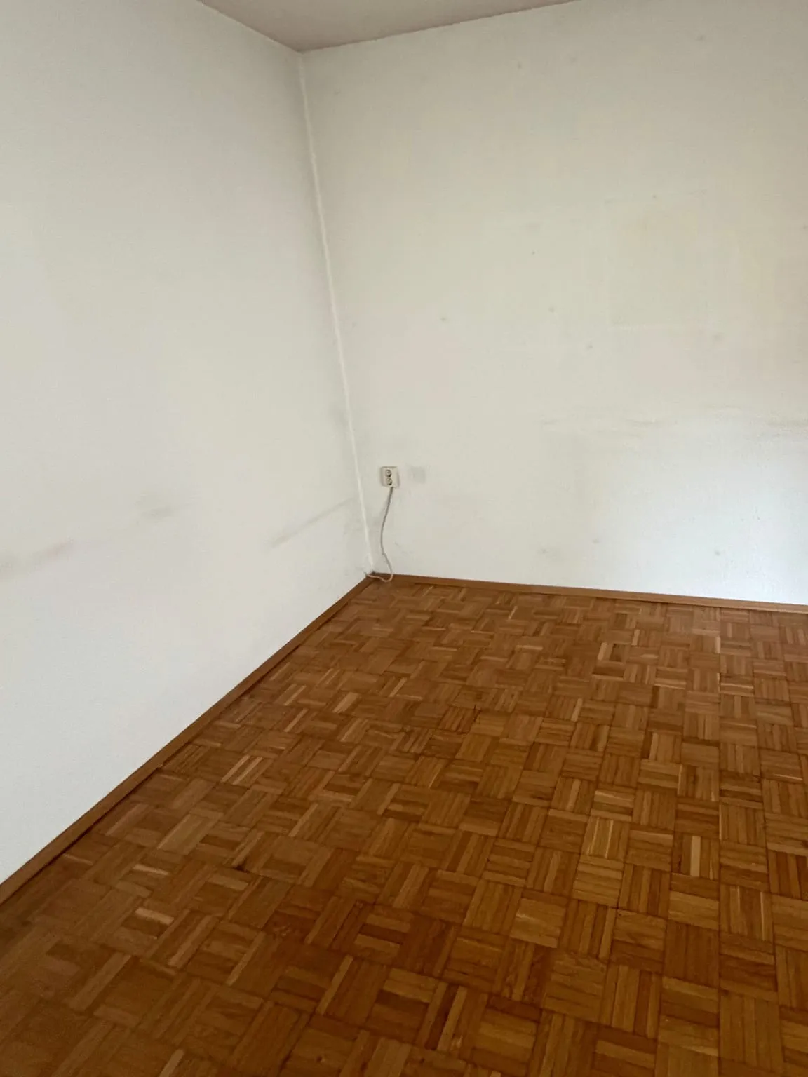 Dieselbe Zimmerecke leer, mit weißen Wänden, einer Steckdose an der Rückwand und freiem Parkettboden