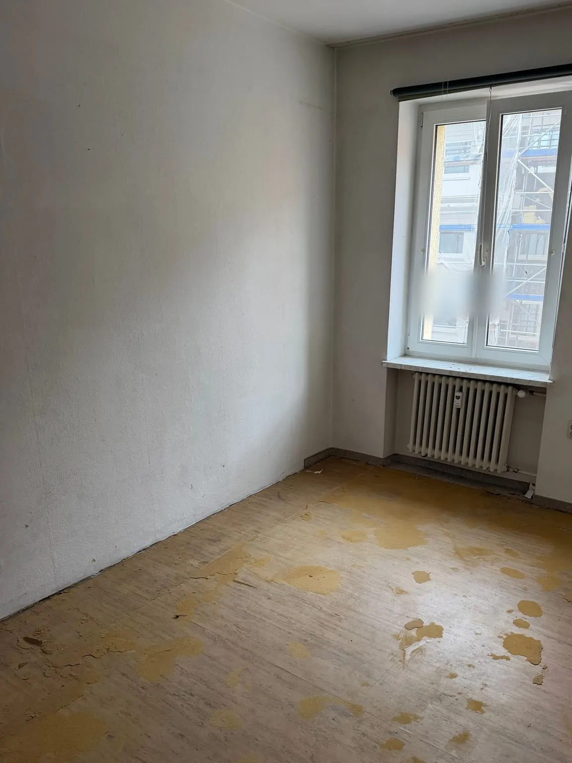 Dasselbe Zimmer leer, mit kahler Wand, Fenster über dem Heizkörper und Holzboden mit Kleberesten des entfernten Teppichs