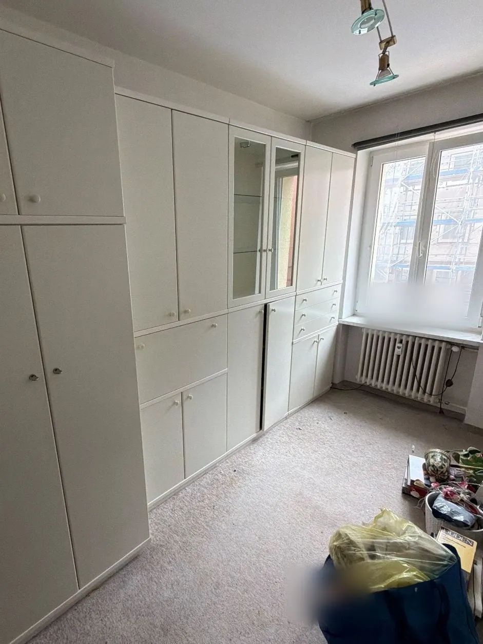 Zimmer mit weißer Schrankwand samt Vitrine entlang der Wand, Teppichboden und einer Tasche mit Hausrat vor dem Fenster